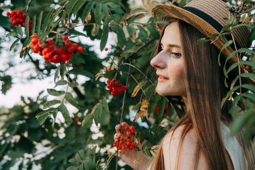A beautiful blonde girl in a hat with long hair, on a walk in the autumn season. Portrait of a woman at a rowan tree.