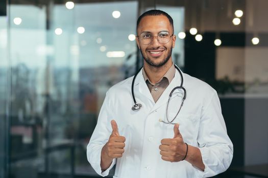 Successful african american doctor looking at camera and smiling holding thumbs up, man in medical coat and glasses with stethoscope working inside modern clinic office.