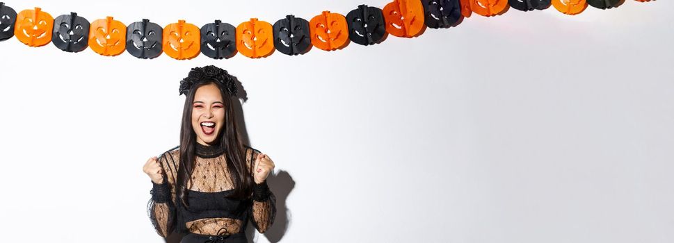 Happy asian woman enjoying halloween, wearing wicked witch costume and rejoicing against pumpkin streamers decoration.