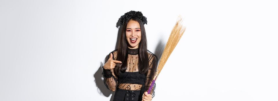 Portrait of attractive woman in witch costume celebrating halloween, pointing at broom, standing over white background.