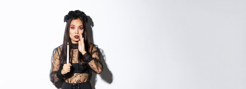 Tensed and worried asian woman in witch costume holding lit candle and whispering, standing over white background.