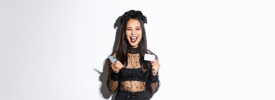 Happy laughing asian girl in halloween costume looking upbeat, holding smartphone and credit card, standing over white background.