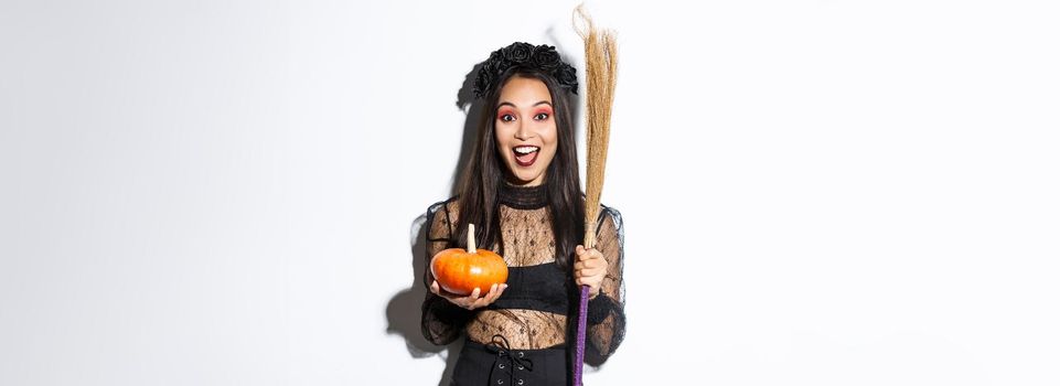 Excited attractive asian girl celebrating halloween, wearing witch costume, holding pumpkin and broom, going trick or treat, standing over white background.