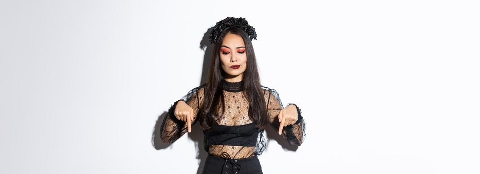 Hesitant and serious-looking asian female in magician costume, dressed-up as evil witch for halloween, looking and pointing fingers down with disbelief, standing over white background.