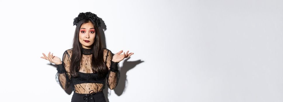 Image of clueless and unbothered asian girl looking confused, shrugging while standing over white background in halloween costume, dressed-up like evil witch on party.