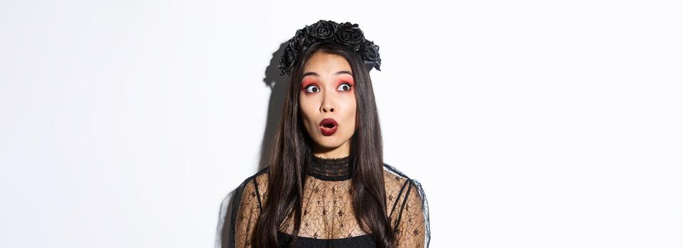 Close-up of surprised asian woman in witch costume looking at upper left corner and open mouth wondered, standing over white background.