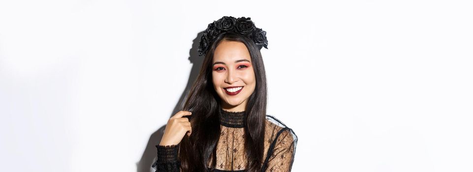 Close-up of beautiful elegant asian woman in black wreath and gothic lace dress smiling, standing over white background, dressed-up for halloween party.