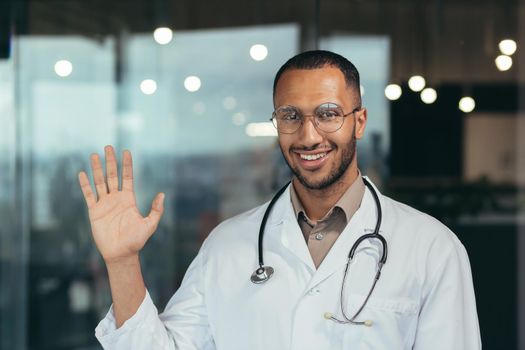 Young african american doctor man wearing glasses wearing medical coat saying hello happy and smiling friendly welcoming gesture doctor working inside clinic.