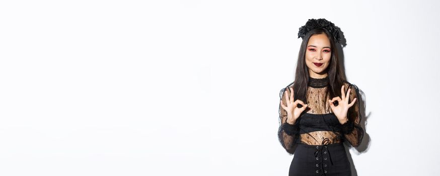 Image of smiling asian cute witch wearing black gothic dress and wreath showing okay gestures in approval, praise something, standing in halloween costume against white background.