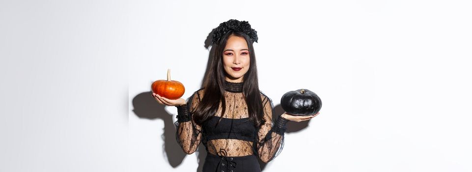 Pleased smiling asian woman celebrating halloween, wearing witch costume, holding pumpkins, standing over white background.