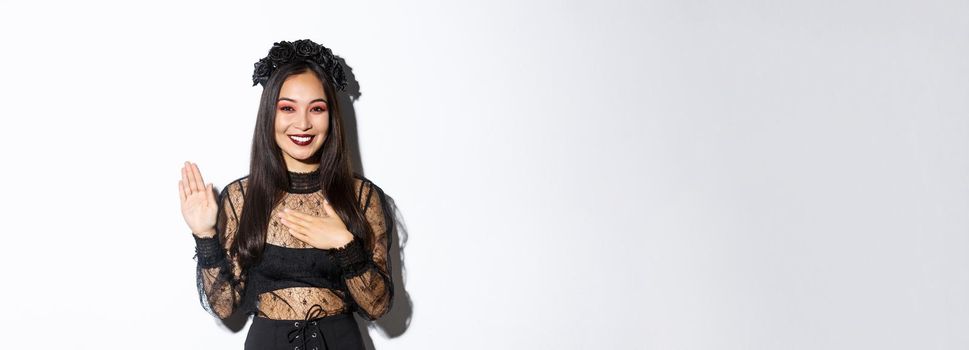 Image of cute sincere asian girl in halloween costume making promise, holding one hand on heart while swearing, being honest, standing over white background.