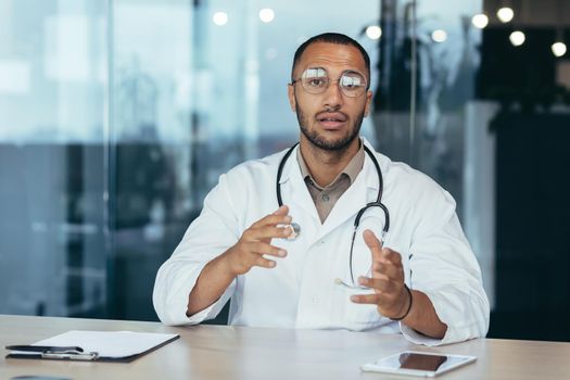 Serious and focused doctor in medical coat and stethoscope talking on video call looking at camera and consulting patient, man working inside clinic office.