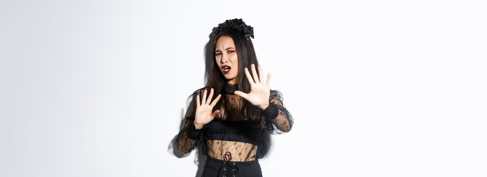 Image of bothered and annoyed asian woman in elegant gothic dress raising hands defensive, grimacing from camera flesh, asking to stop taking pictures of her, standing white background.