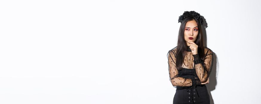 Thoughtful elegant evil witch in black lace dress, gothic wreath and makeup looking upper left corner. Woman making plans for halloween party, thinking over white background.