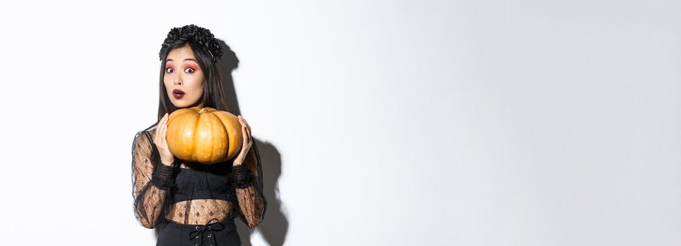 Portrait of woman lifting heavy pumpkin, getting ready for halloween, wearing witch costume, standing over white background.