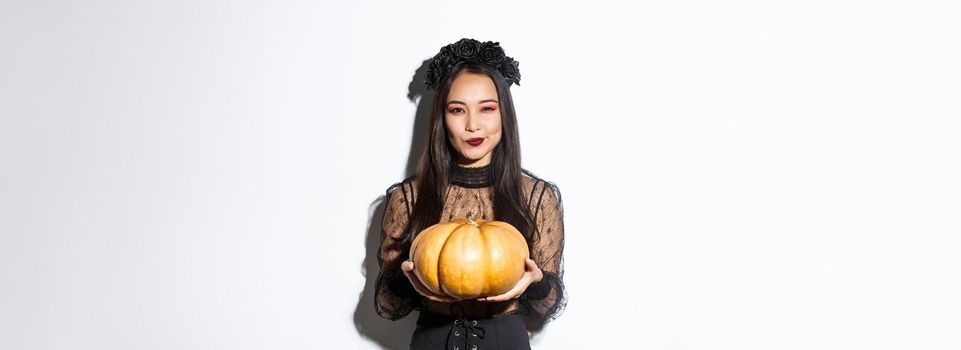 Image of cunning asian woman in black dress, impersonating evil witch on halloween, holding big pumpkin, standing over white background.