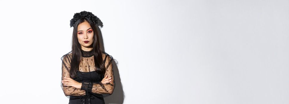 Skeptical and unamused asian woman dressed in halloween costume looking disappointed at camera, cross arms chest. Female in black gothic dress and wreath judging someone.