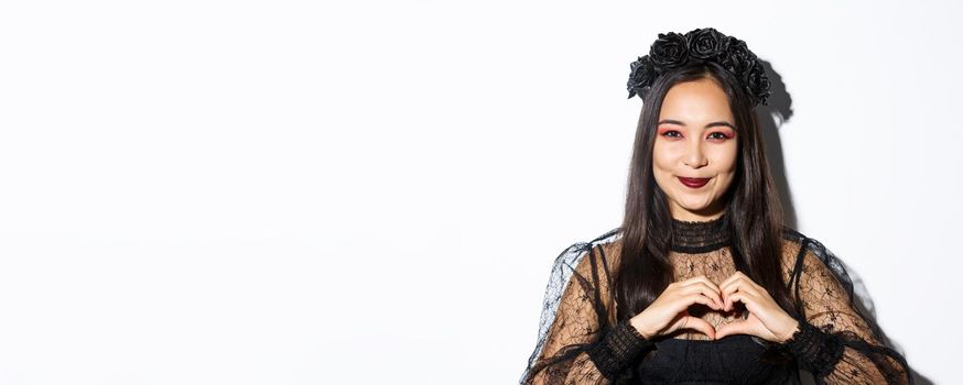 Close-up of smiling pretty asian woman in witch costume and black wreath showing heart gesture, loving halloween holiday, standing over white background.