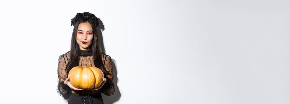 Image of cunning asian woman in black dress, impersonating evil witch on halloween, holding big pumpkin, standing over white background.