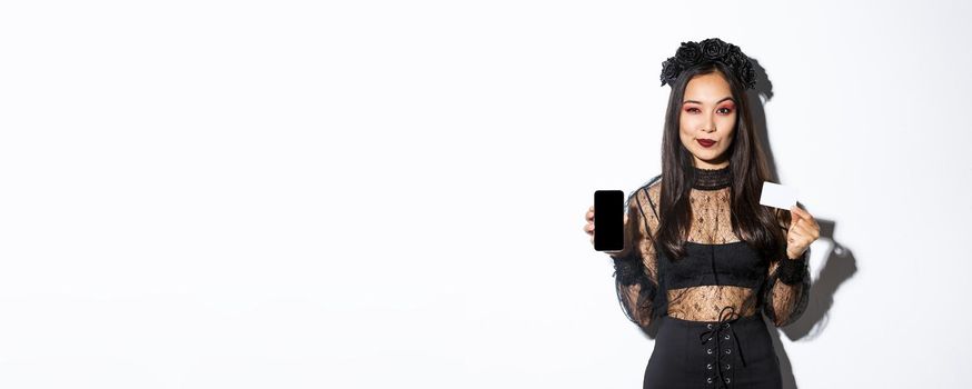 Sassy elegant young witch in gothic lace dress and black wreath showing you credit card and mobile phone screen, standing over white background.