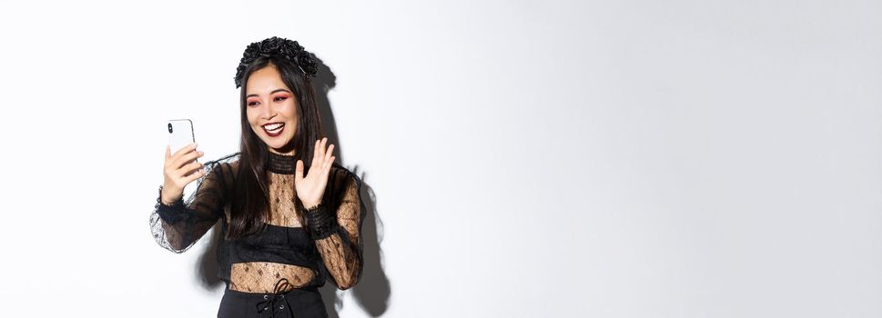 Portrait of beautiful and stylish asian woman in gothic lace dress saying hi, waving hand at smartphone camera during video call, standing over white background.