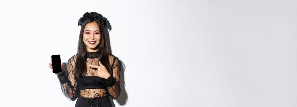 Happy beautiful asian girl in witch costume pointing finger at smartphone screen with pleased smile, showing halloween announcement, white background.