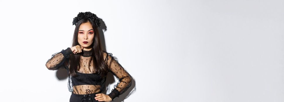 Image of beautiful gothic asian woman in black lace dress, threaten someone, make cutting throat gesture and looking serious at camera, celebrating halloween, standing over white background.