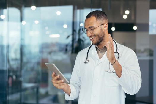 Cheerful hispanic doctor in medical white coat and stethoscope using tablet computer for video call and online consultation to patients, man working inside clinic office building.