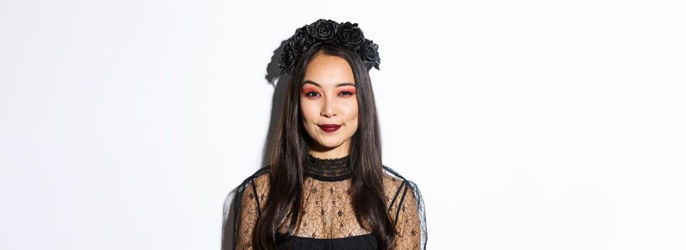 Image of smiling devious young witch looking at camera with cunning smirk, standing over white background. Woman in witch costume celebrating halloween.