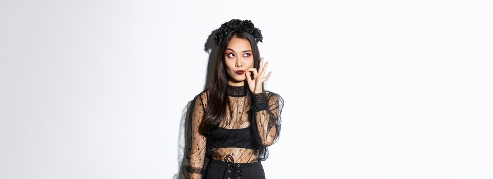 Young gothic woman in black lace dress, dressed-up like witch for halloween party, making seal on lips, zipping mouth as making promise keep secret, standing over white background.