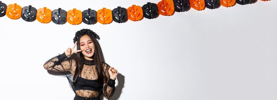 Carefree smiling asian woman in witch costume enjoying halloween party, dancing and showing peace gesture, standing over white background with pumpkin decoration.