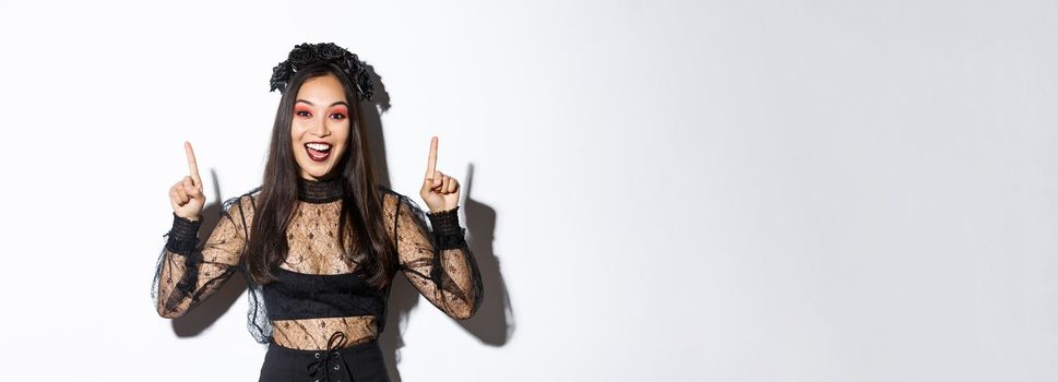 Excited smiling woman celebrating halloween in black gothic dress and wreat. Asian girl wearing witch costume and pointing fingers up, standing white background.