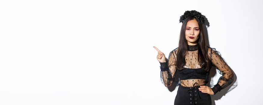 Image of disappointed and skeptical asian woman in witch costume complaining on something, pointing upper left corner and grimacing dissatisfied, standing over white background in halloween dress.