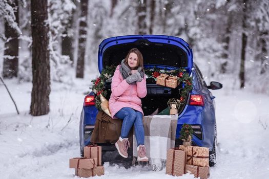 A woman in winter clothes drinks a hot drink sitting in a car decorated in a New Year's way. Traveling by car through the forest. A trip before Christmas.