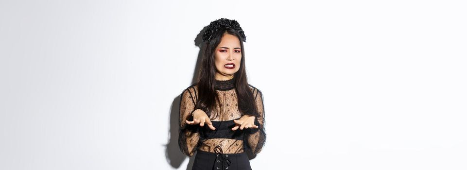 Image of funny asian woman in halloween witch costume looking disgusted, refuse from something nasty or creepy, standing over white background, express aversion.