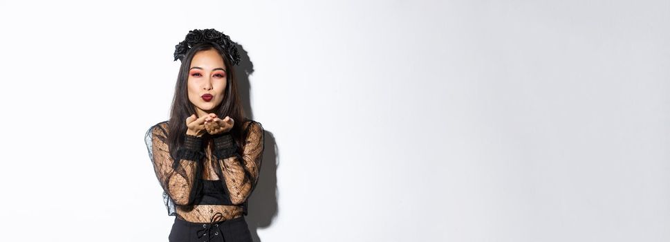 Image of sexy asian woman sending air kiss at camera, ready for halloween party, wearing gothic dress and wreath, celebrating all saints day, standing over white background.