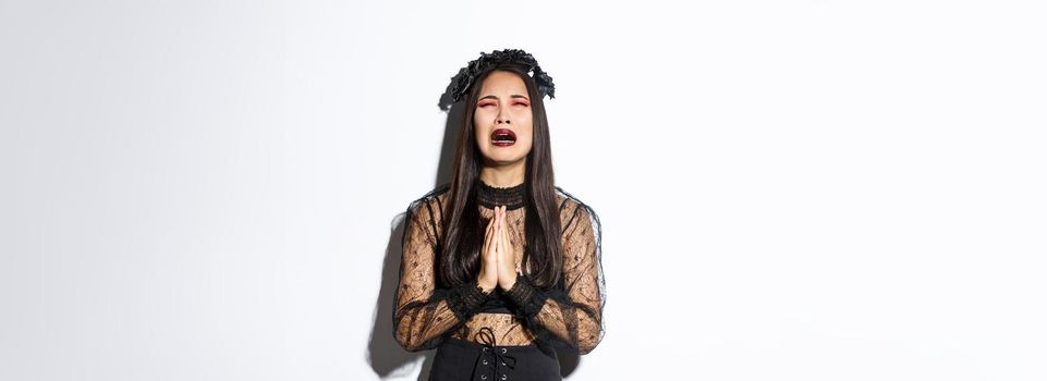 Miserable young asian woman in trouble pleading god, crying and begging for help, wearing halloween gothic dress and wreath, supplicating over white background.