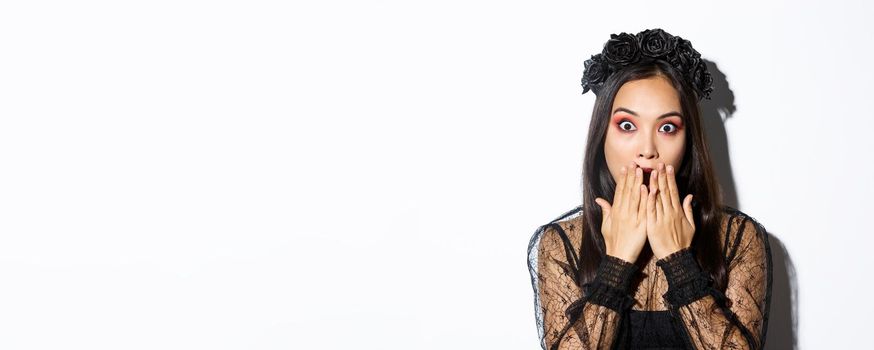 Close-up of shocked asian woman in gothic lace dress and wreath gasping, cover mouth and stare at camera amazed, standing over white background.