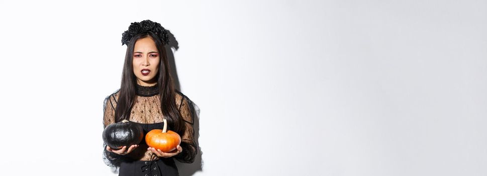 Image of grimacing angry witch throwing pumpkins, girl celebrating halloween, standing over white background.