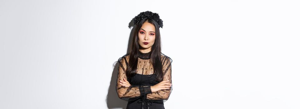 Skeptical and unamused asian woman dressed in halloween costume looking disappointed at camera, cross arms chest. Female in black gothic dress and wreath judging someone.