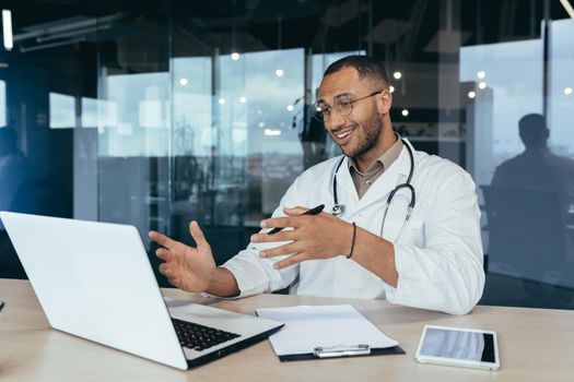 Cheerful african american doctor talking on video call, man using laptop for remote online consultation of patients, doctor working inside modern clinic in office.