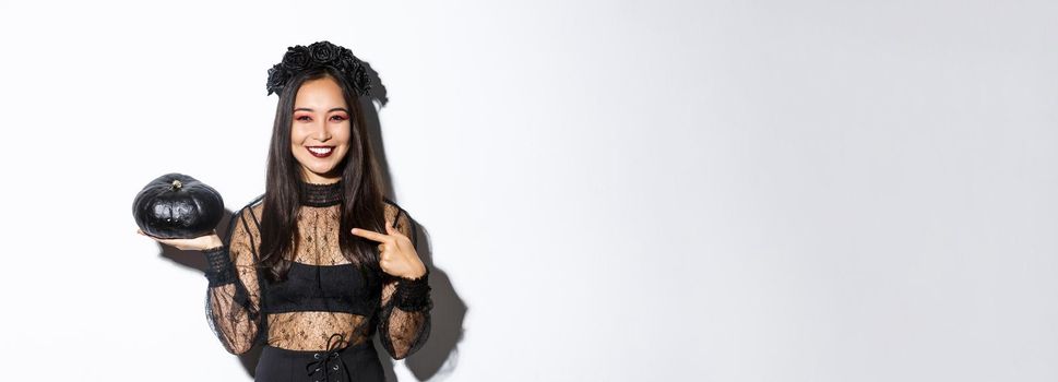 Happy smiling asian woman enjoying halloween celebration, wearing witch costume and pointing finger at black pumpkin, standing over white background.