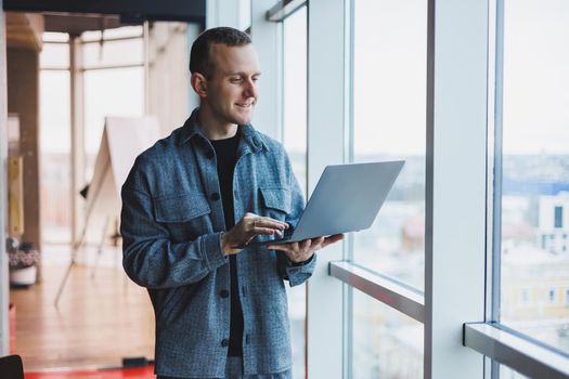 Portrait of a smart intelligent male manager, he is holding a laptop for research in the office, a business man in elegant clothes playing on the touchpad. Standing in the office by a large window