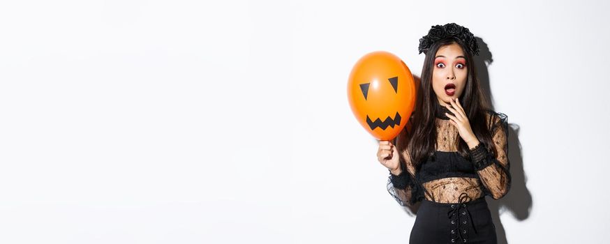 Portrait of surprised asian woman in halloween costume, dressed-up as witch, holding orange balloon with scary face.