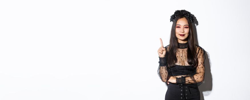 Creative young woman in witch costume smiling pleased as have great idea, raising finger to say suggestion. Female asian dressed-up like widow or mysterious magician, white background.