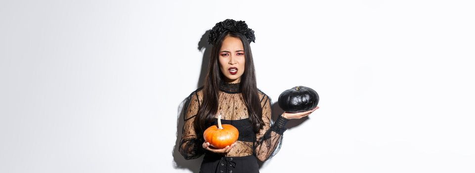 Portrait of disappointed frowning girl in witch costume, holding pumpkins and complaining, grimacing while standing over white background.