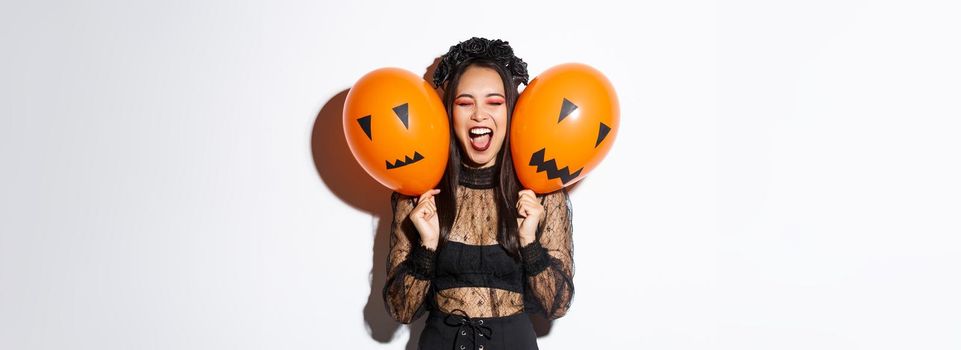 Image of asian girl in evil witch costume holding two orange balloons with scary faces, celebrating halloween, standing over white background.