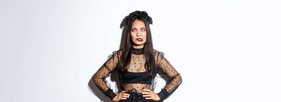Annoyed asian woman in witch costume rolling eyes and looking pressured. Irritated girl hate celebrating halloween, looking pissed-off, wearing gothic black lace dress and wreath.