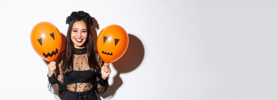 Image of cheerful asian woman in witch costume celebrating halloween, holding balloons with scary faces, standing over white background.