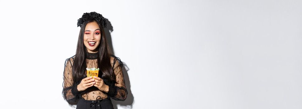 Cheeky smiling girl in witch costume, celebrating halloween, going trick or treat in gothic dress, showing tongue and holding sweets.